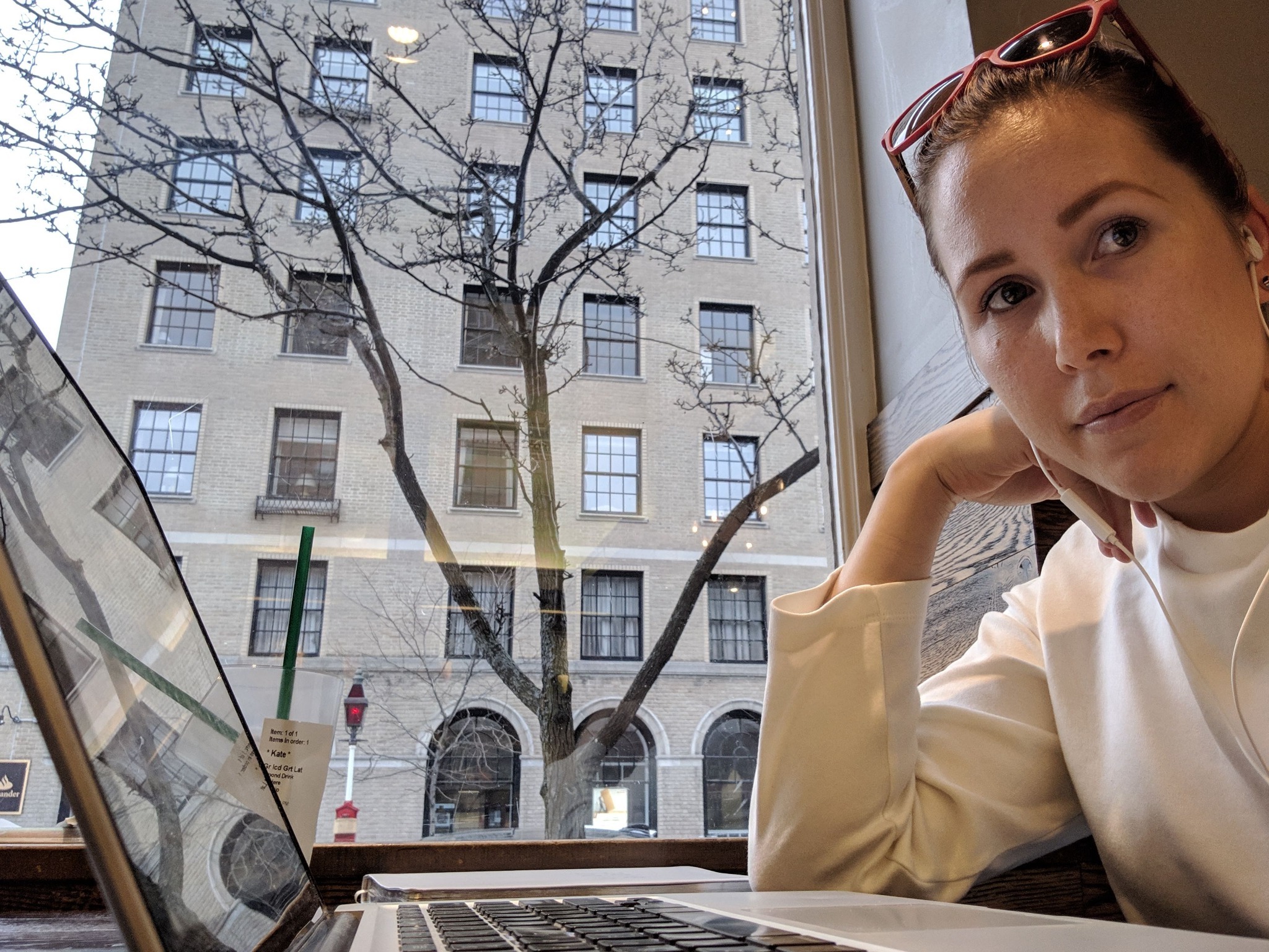 Young woman wearing white with red sunglasses on head, sitting near window with laptop open