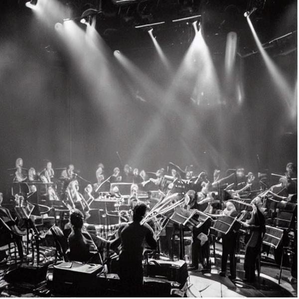 black-and-white photo of a symphony orchestra from the audience's perspective with 6 intersecting spotlights overhead