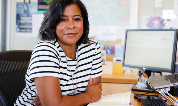 African American woman with black hair wearing black-and-white striped shirt leans on her forearms at a desk, facing the camera but sitting in front of a monitor. She is just slightly smiling. The slightly blurred background may show artwork and books.