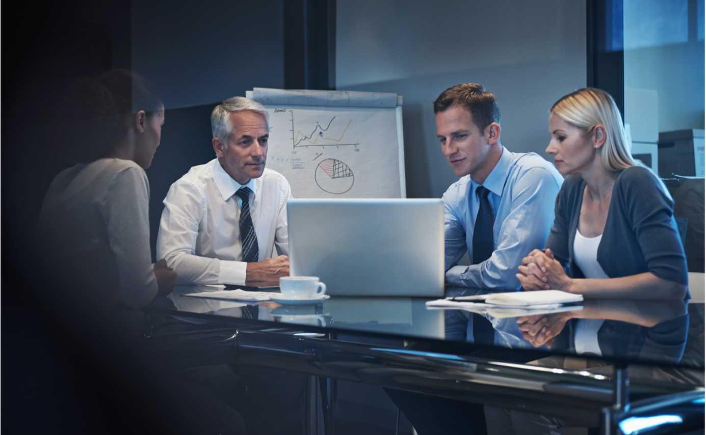 Team of 4 collaborating in an office around a desk with laptop and easel with charts Team of 4 collaborating in an office around a desk with laptop and easel with charts