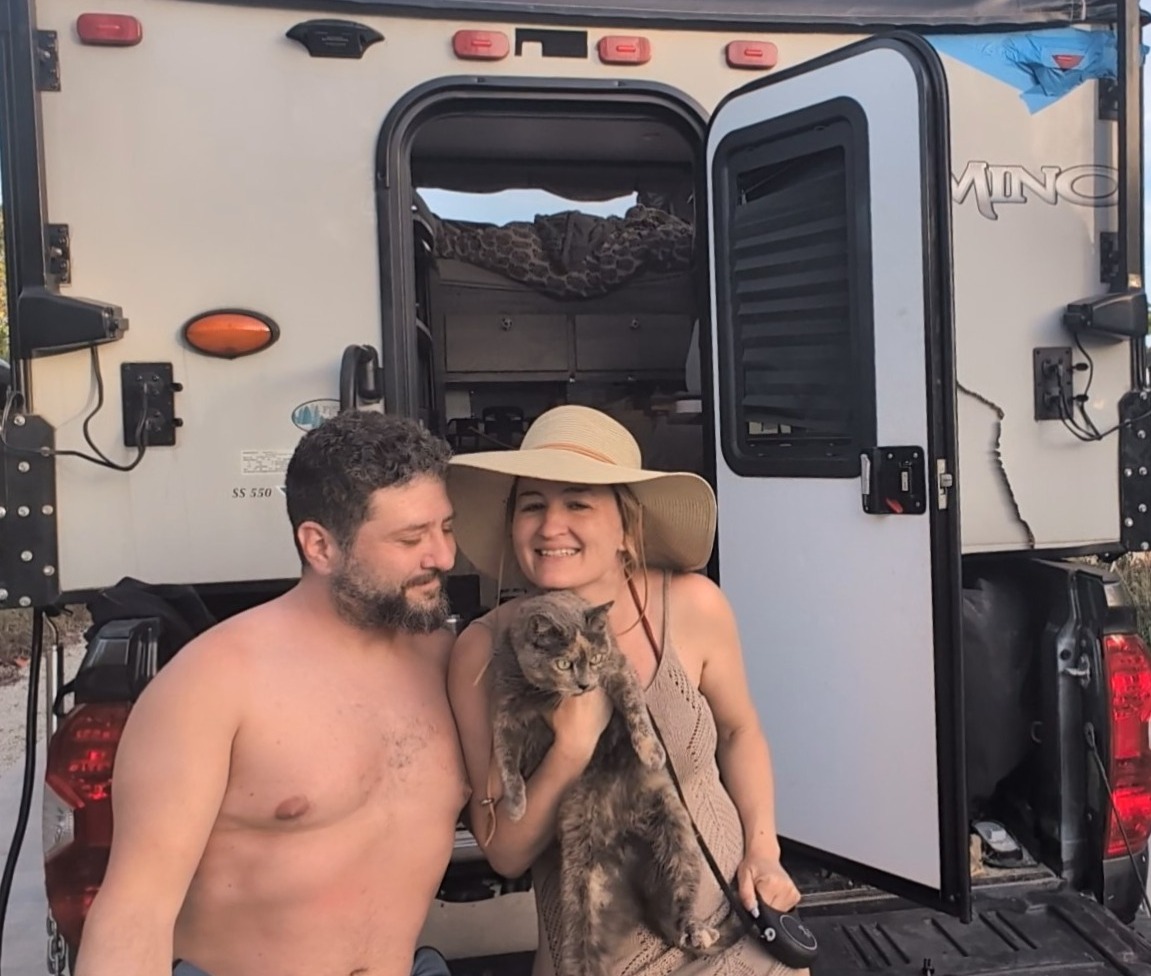 A middle-aged couple in beachwear sitting on the bumper of a truck camper holding a tortishell cat on a leasht
