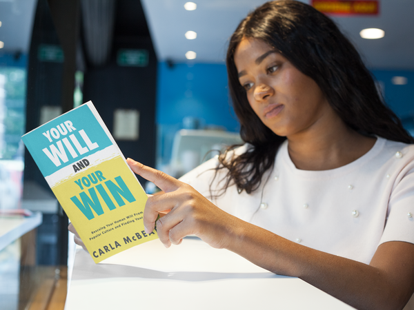 Young Black girl in white shirt reading a paperback titled Your Will and Your Win