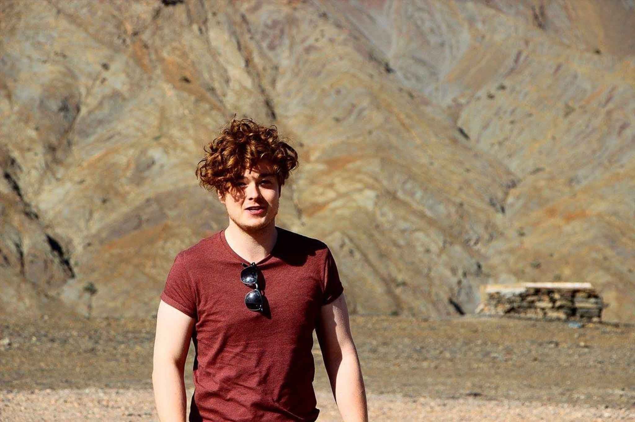 A man with red curly hair walking toward the camera with sandy mountains behind him A man with red curly hair walking toward the camera with sandy mountains behind him