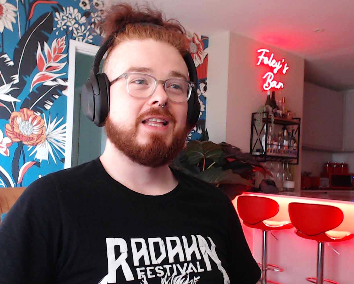 Tom Foley, games journalist, sitting in a chair while talking with a red bar sign and stools in the background Tom Foley, games journalist, sitting in a chair while talking with a red bar sign and stools in the background