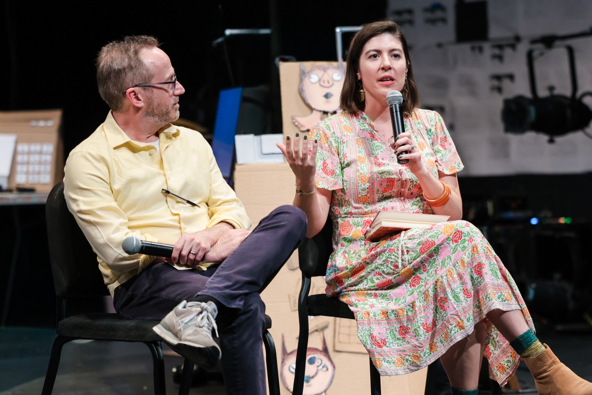 Amelia on microphone explaining in a colorful dress, seated next to a white man in a yellow button-down shirt