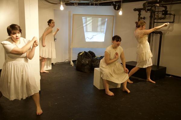 Four women dressed in white nightgowns hold statuesque poses in a white walled room