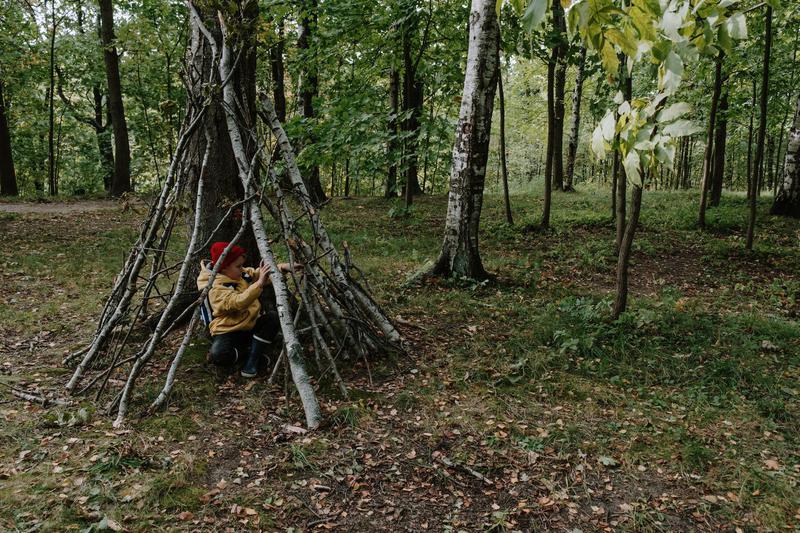 This is a photo of a person in a red knit cap and yellow jacket kneeling inside a fortress made of sticks. This person is inside this stick-fortress in the middle of the woods.