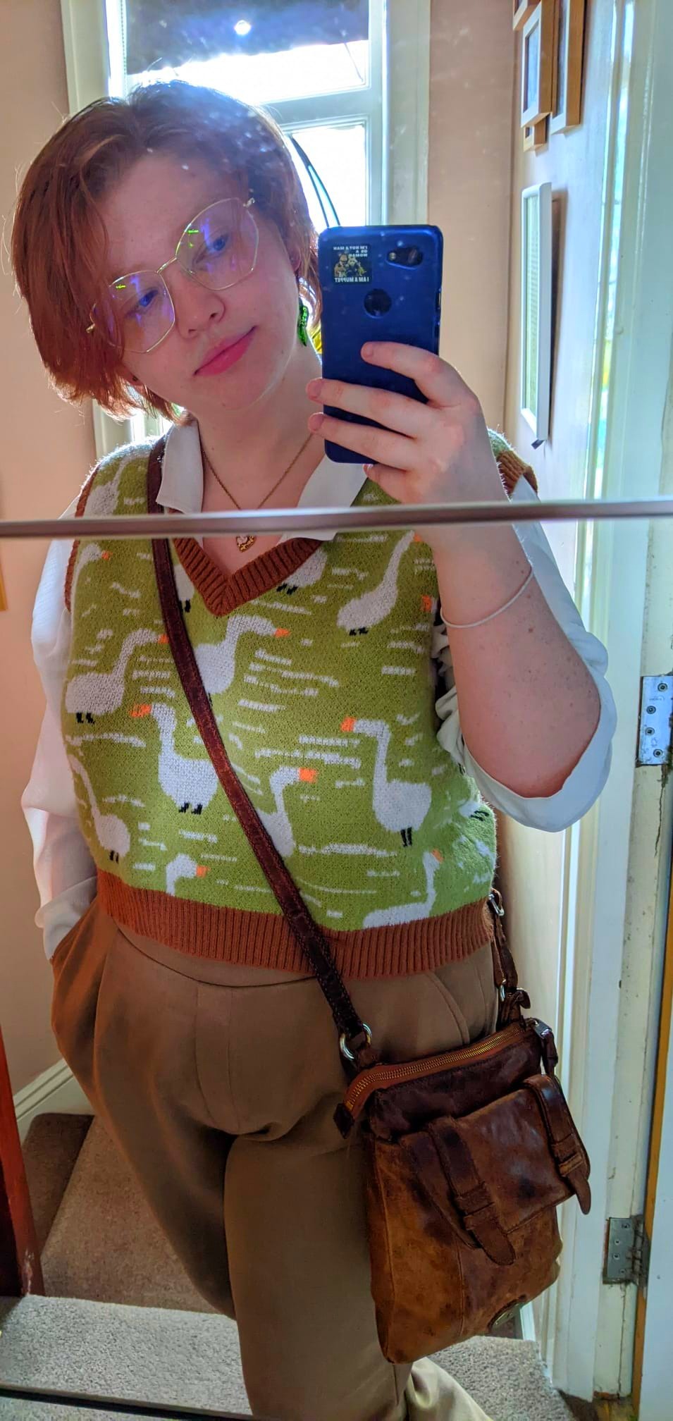 A selfie taken in a mirror of a small red-haired guy in a fabulous goose vest, white shirt and brown trousers with a leather satchel.