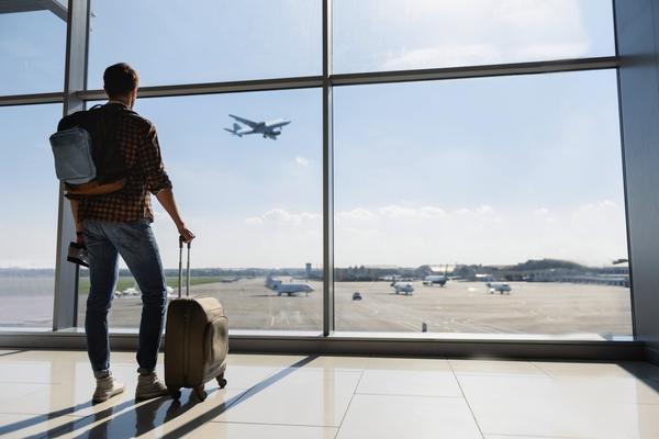 Young man is standing near window at the airport and watching plane before departure. He is standing and carrying luggage. Focus on his back