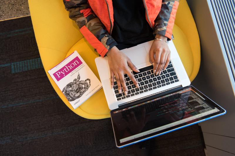 A photo of a Python developer sitting at a desk, coding on a computer. A book titled 'Python Development' is visible on the desk next to the computer