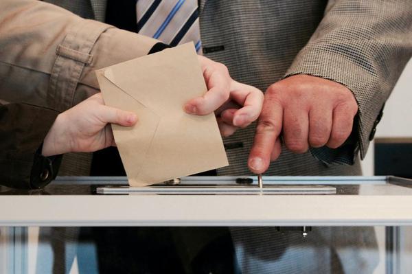 Man pointing to a ballot box slot as another casts a ballot