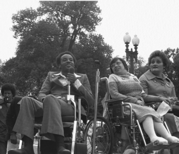 Brad Lomax is seen speaking with a mic in a wheelchair. Judy Heumann is seen in a wheelchair beside him, with another white woman beside her. To their left, a man is seen smoking, with trees in the background.
