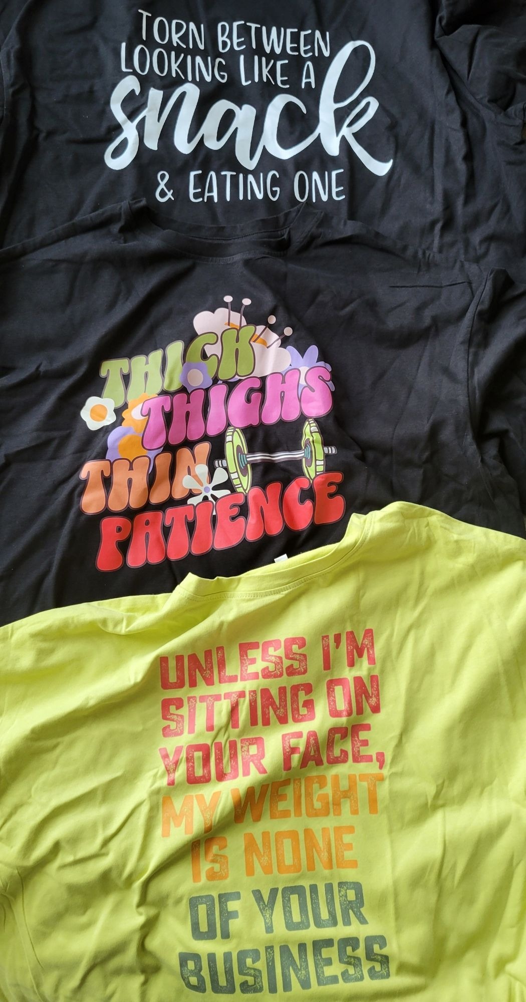 3 rumpled crop tops are seen.  The black one from the top states this in white text: TORN BETWEEN LOOKING LIKE A SNACK& EATING ONE  The black one in the middle states this in green, pink, orange, and red text: THICK THIGHS THIN PATIENCE  The green one below states this in red, orange and blue: UNLESS I'M SITTING ON YOUR FACE, MY WEIGHT IS NONE OF YOUR BUSINESS