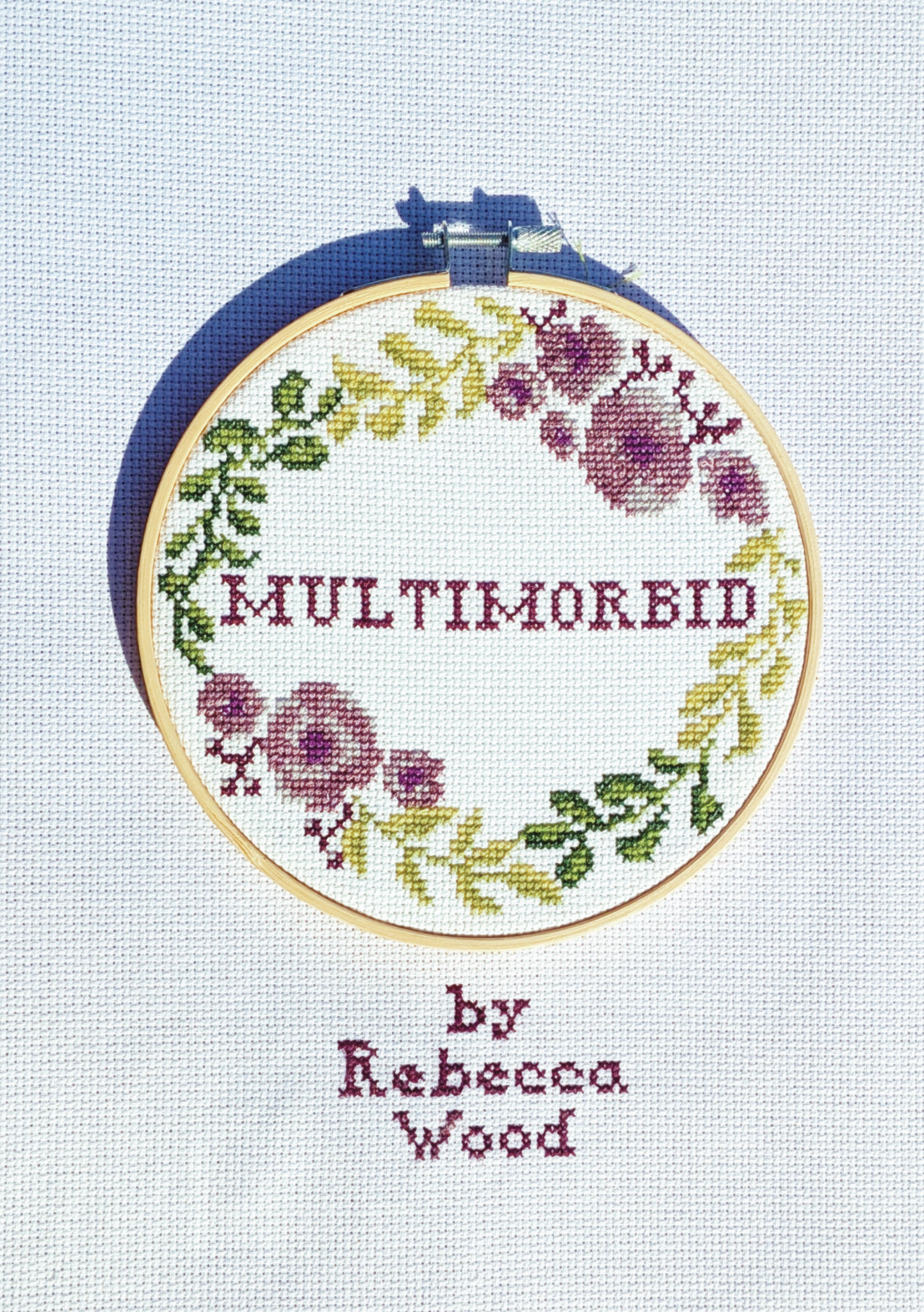 Title: Multimorbid by Rebecca Wood The book's cover is a photograph of an embroidered wreath of purple flowers and green leaves surrounding the title. The piece is held in a wooden hoop set on a background of aida cloth embroidered directly with text reading 'by Rebecca Wood'.