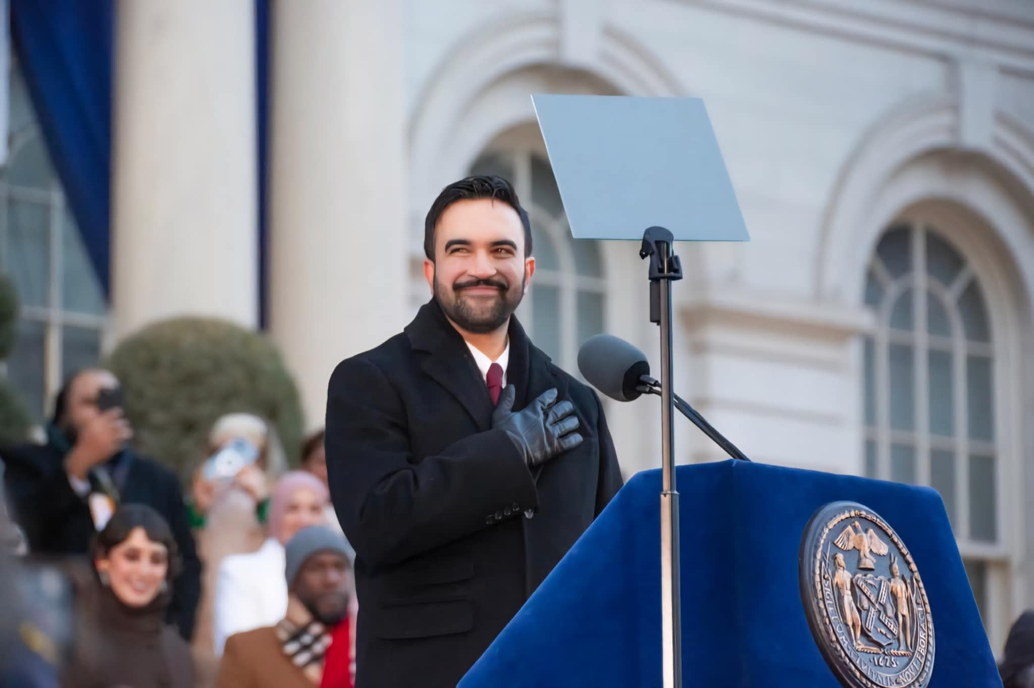 Zohran Mamdani, a brown man, is seen at his inauguration as the new NYC mayor. He is seen outdoors, smiling at an ornate blue lectern. Behind him, Rama Duwaji, his wife, and Jumaane D. Williams, from the NYC Office of the Public Advocate, are seen, among other members of the public of a variety of races, genders, etc.