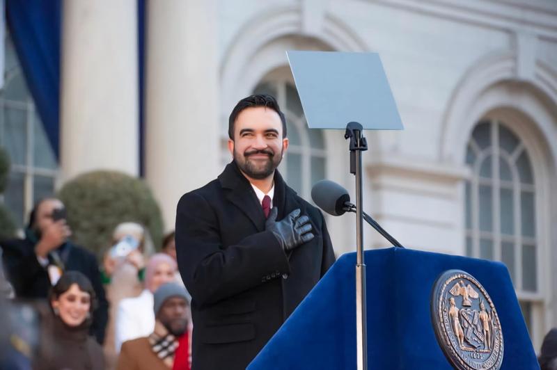 Zohran Mamdani, a brown man, is seen at his inauguration as the new NYC mayor. He is seen outdoors, smiling at an ornate blue lectern. Behind him, Rama Duwaji, his wife, and Jumaane D. Williams, from the NYC Office of the Public Advocate, are seen, among other members of the public of a variety of races, genders, etc.