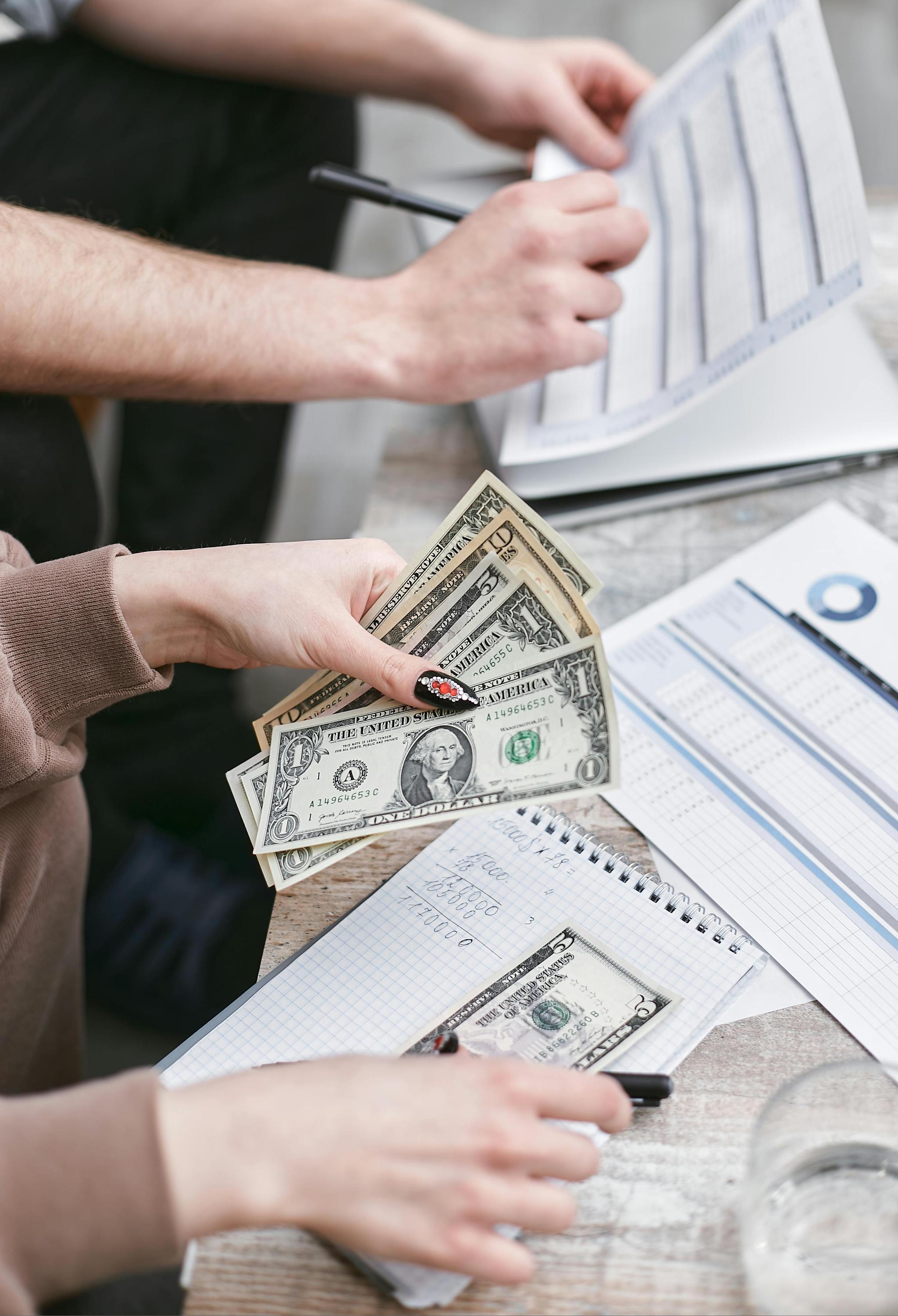A pair of white hands write on paper, while another pair of white hands beside it is counting dollar bills.
