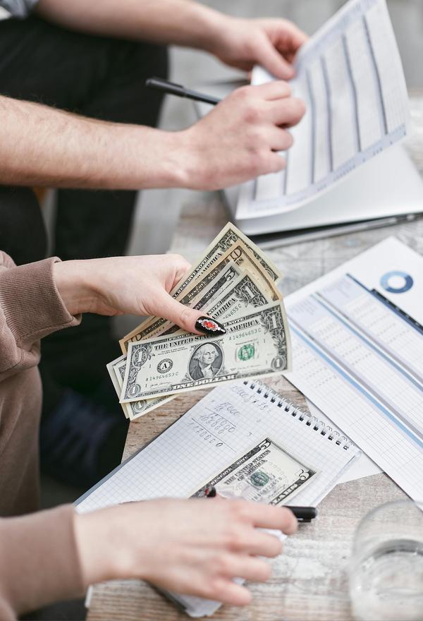 A pair of white hands write on paper, while another pair of white hands beside it is counting dollar bills.