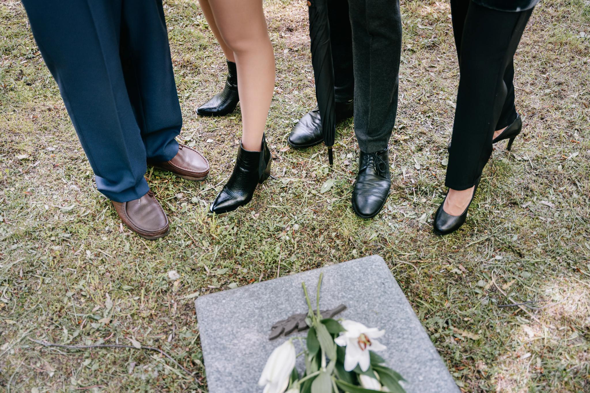 Beside a gray gravestone with flowers, the legs of 4 folx are seen, standing, along with a black umbrella.