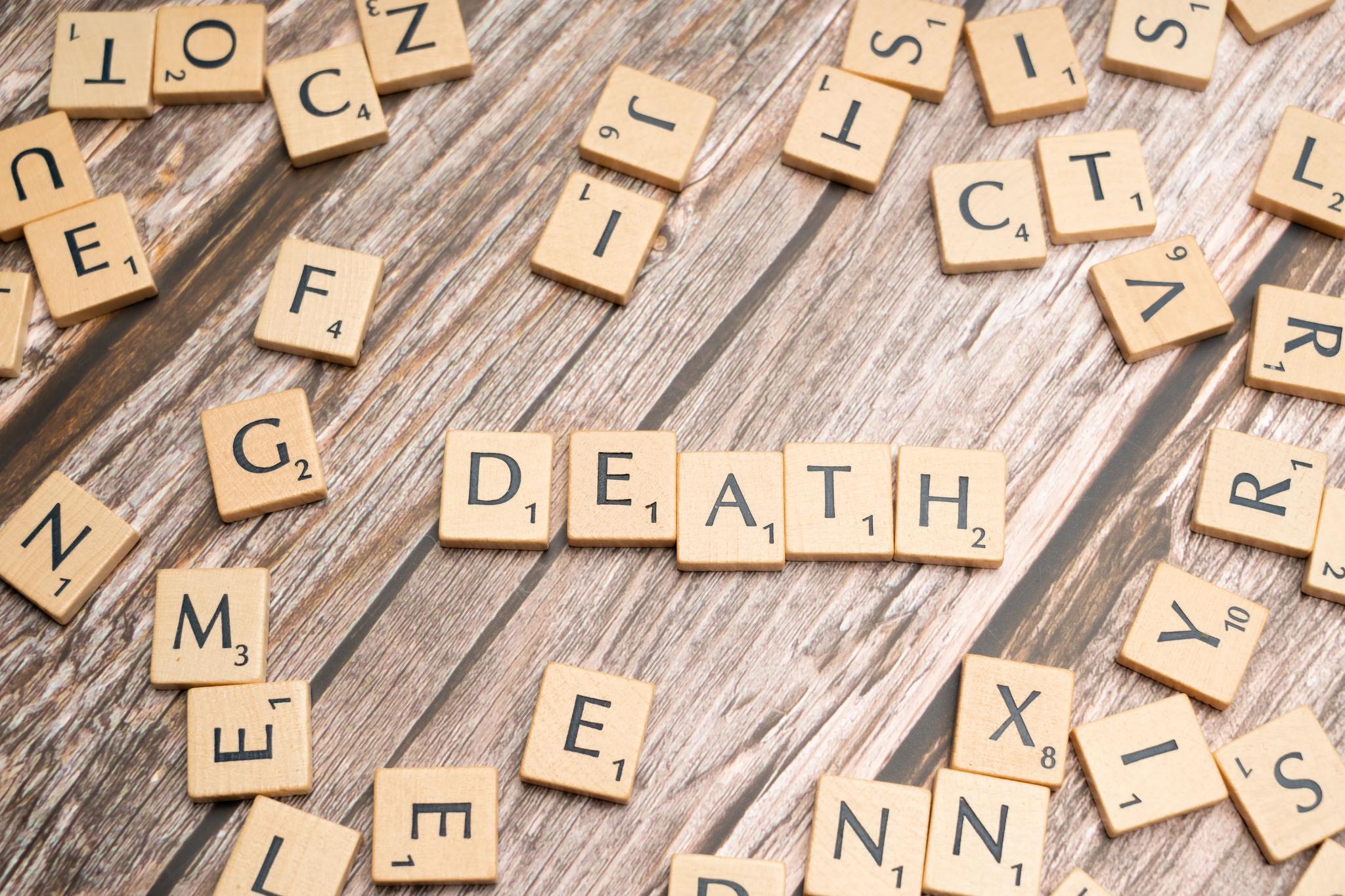 Against a brown background, beige Scrabble letter tiles with black text are seen, with DEATH spelled out in the middle.