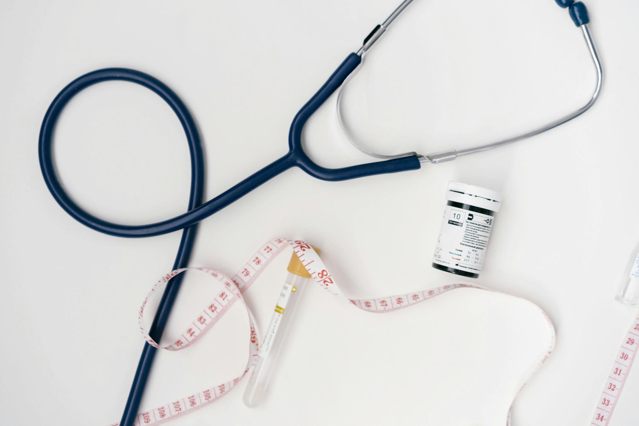 Against a white background, a blue stethoscope is seen, with a measuring tape, a drug container, and a vial.