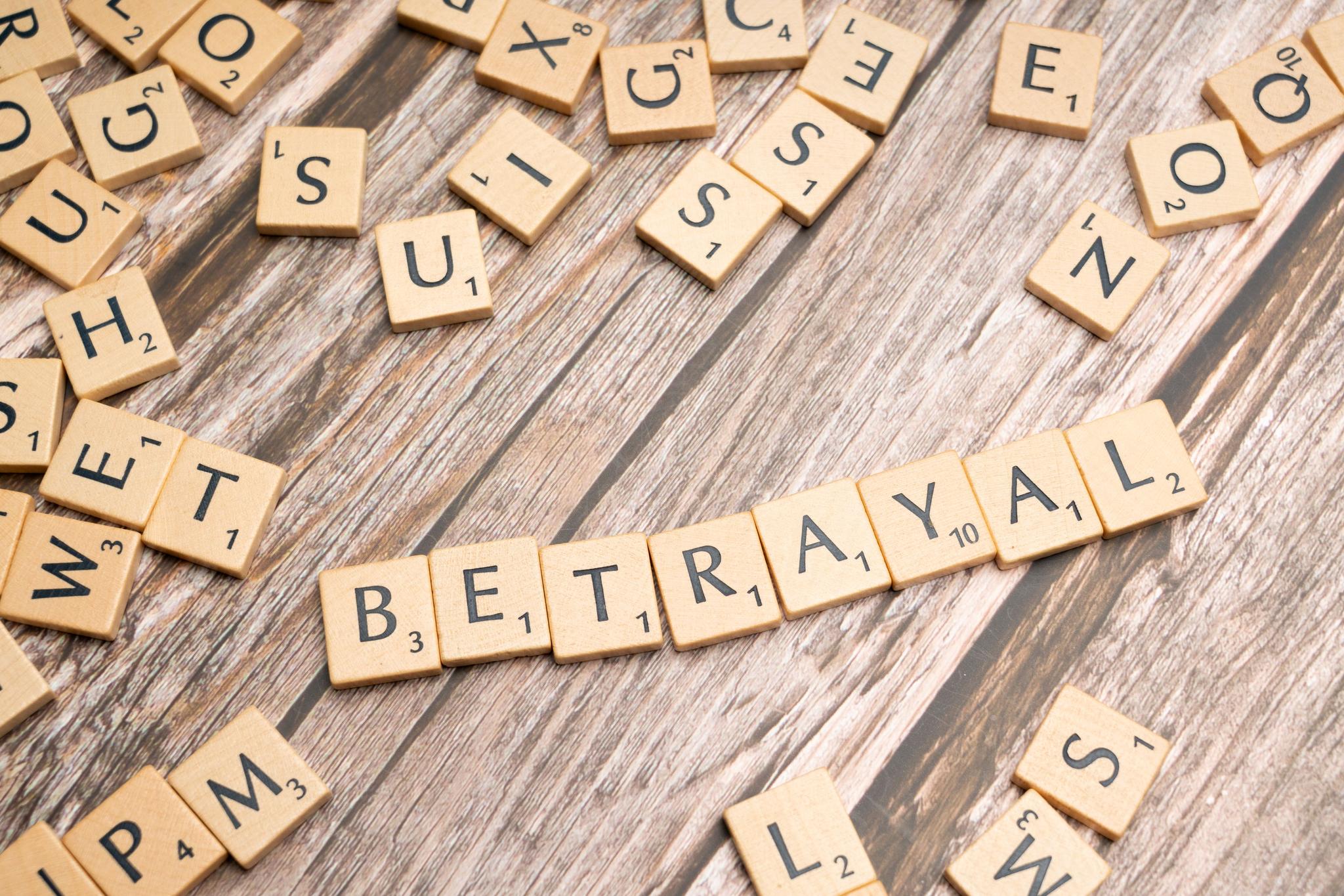 Against a brown background, beige Scrabble letter tiles with black text are seen, with BETRAYAL spelled out in the middle.