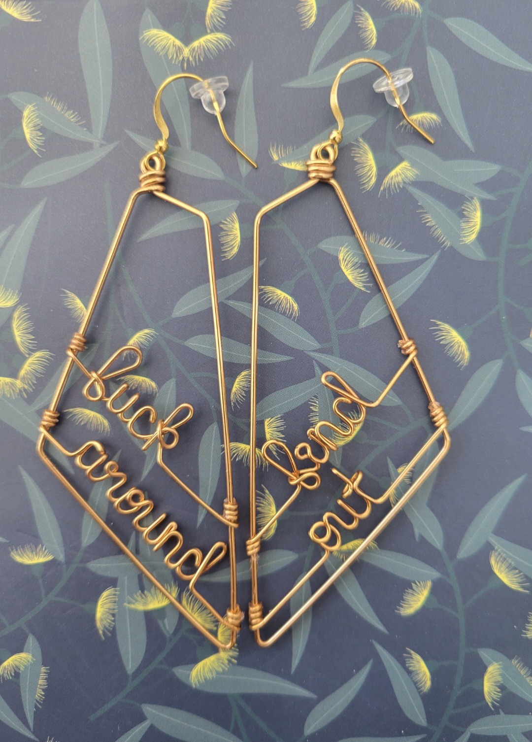 Against a blue, green, and yellow notebook cover, a pair of handmade gold wire earrings from Femmeboyant Designs are seen, which state, "fuck around" and "find out."