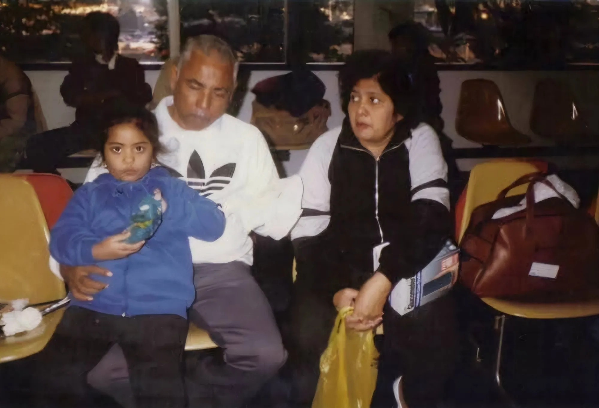 A photograph is seen. Pappy, a grey-haired brown man is seated on an uncomfortable chair, hugging Grims, a black-haired brown woman, with his left hand, while Krystal, a brown girl, sits on his lap, eating a bag of chips, as he hugs her with his right hand.