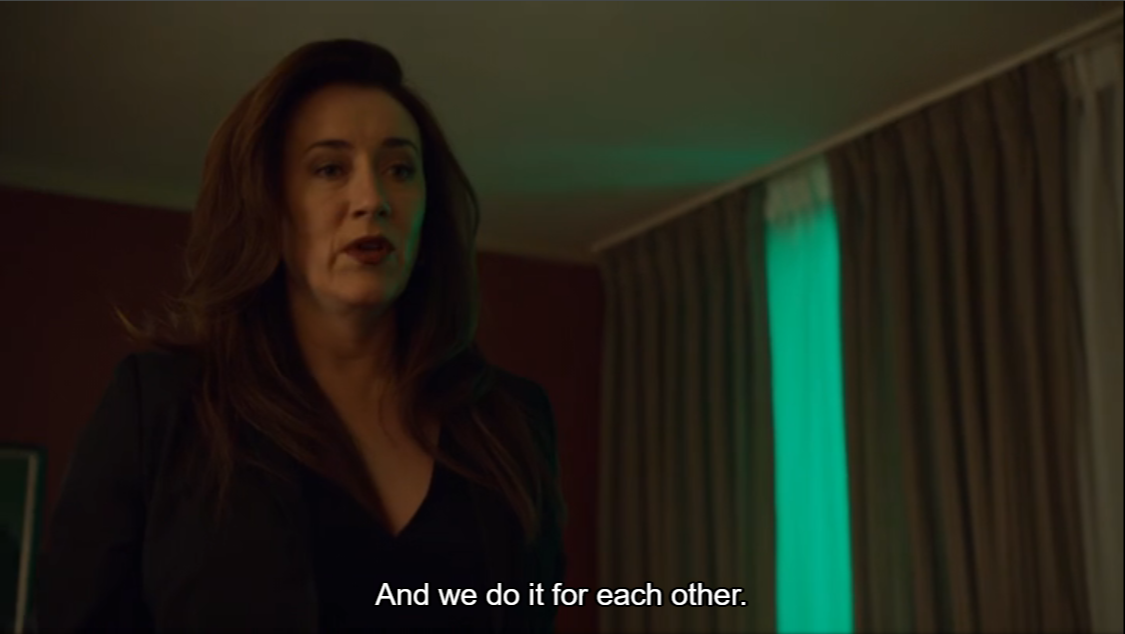 An Orphan Black screenshot is seen. Siobhan, a white woman with auburn hair cascading down her shoulders, is elegantly dressed in a black blazer, with bold lipstick. Captions state: And we do it for each other.