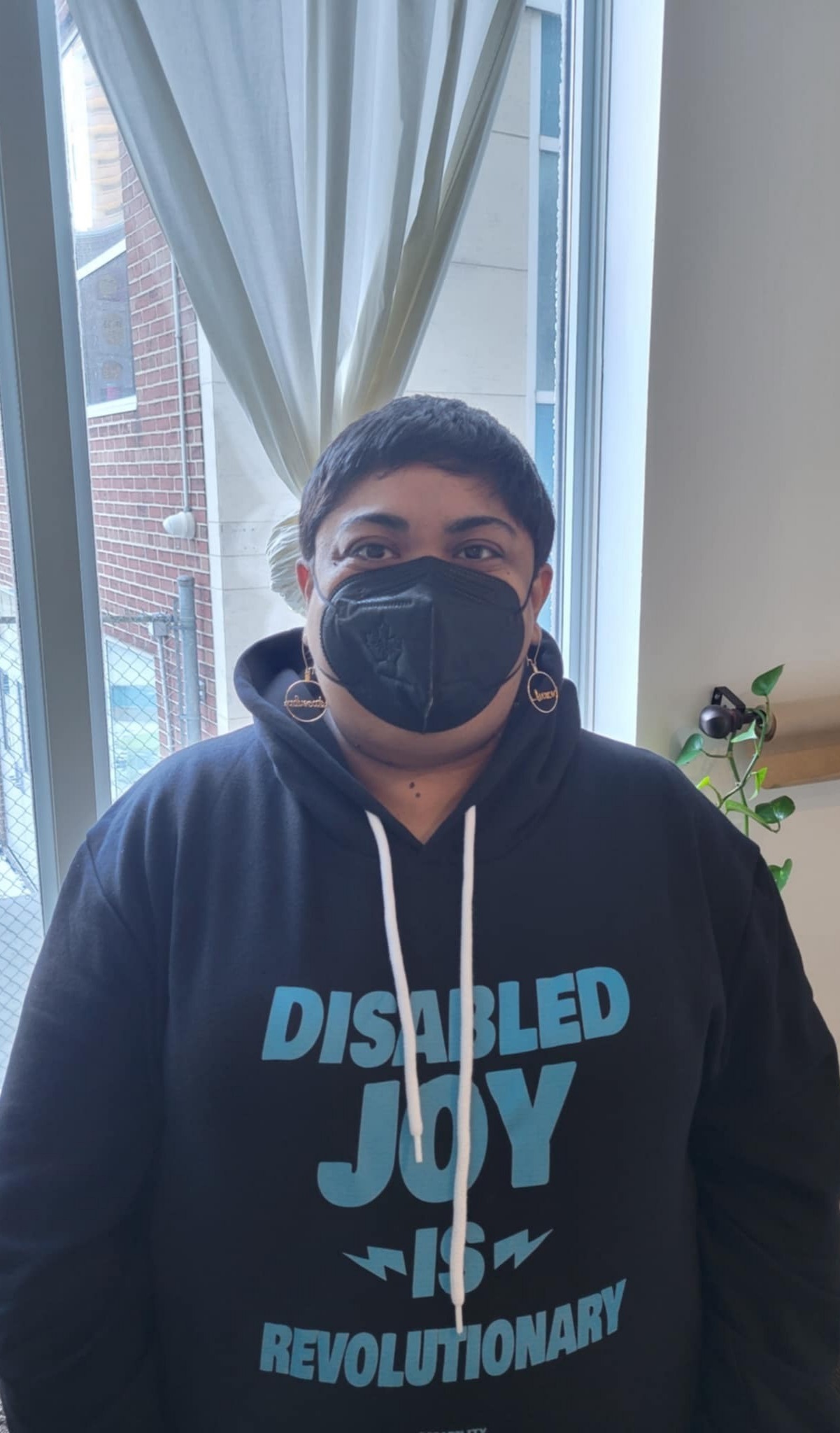 Krystal Kavita Jagoo, a fat brown queer disabled immigrant woman with a pixie cut is seen wearing a black hoodie, a black KN95 mask, and gold earrings, which say "fierce advocate," with a window behind her.