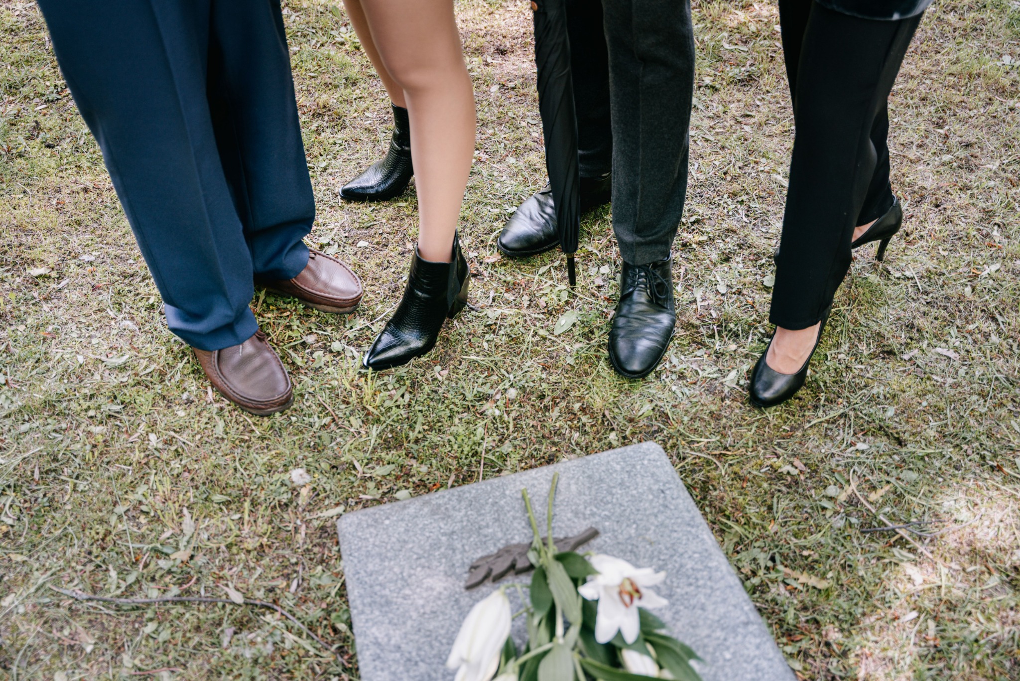 Beside a gray gravestone with flowers, the legs of 4 folx are seen, standing, along with a black umbrella.