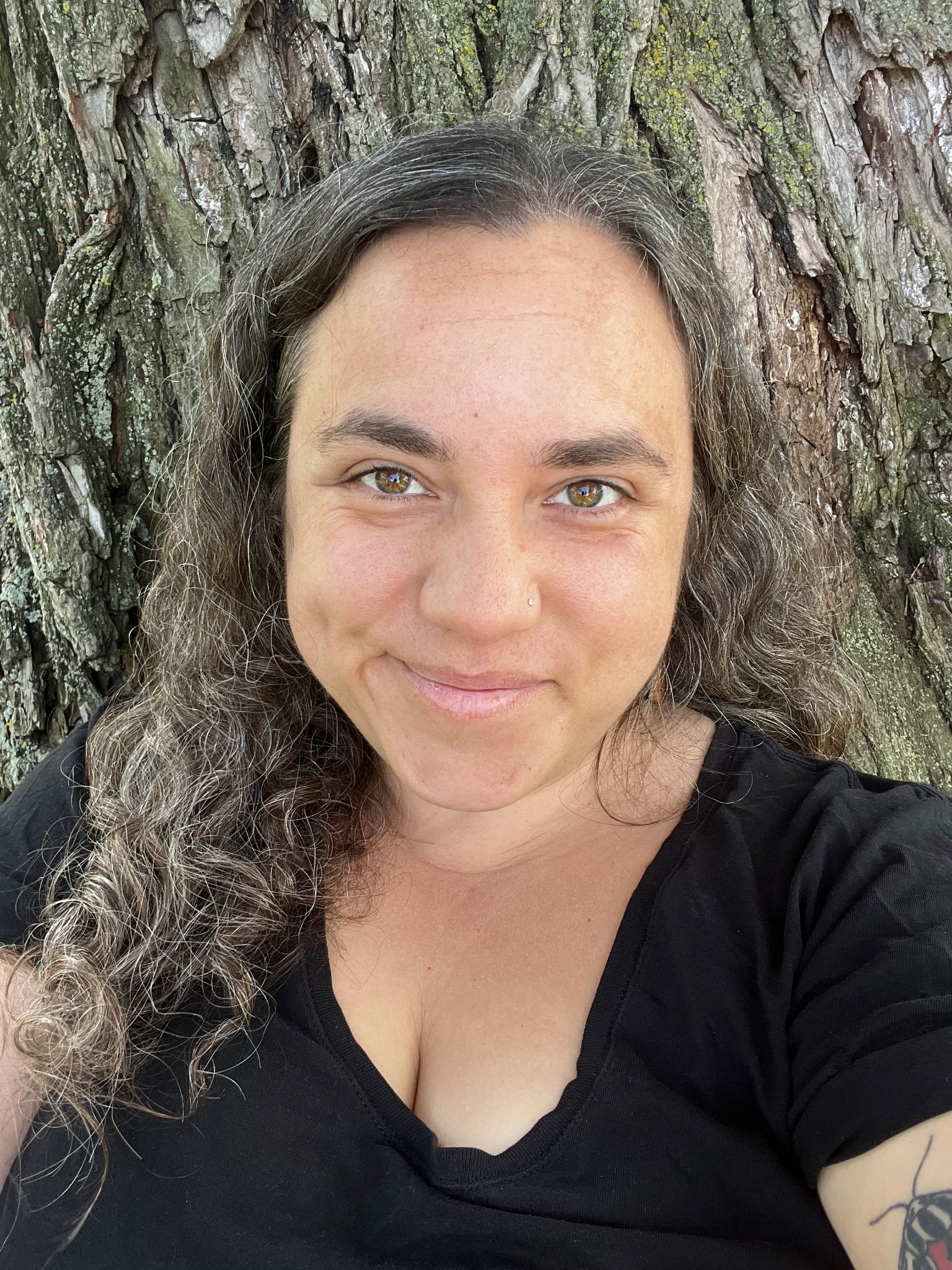 Photo of author Rebecca Wood smiling in front of a linden tree whose bark fully fills the background of the image. Rebecca is a white woman with a round face, long curly dark brown and grey hair, with thick eyebrows, hazel eyes and a sparkling nose stud. She is wearing a black v-neck tshirt and has a virgin tiger moth tattoo on her left arm that is just peeking into frame.