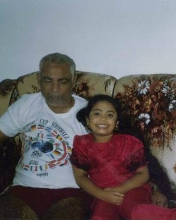 A cropped photo from the author's childhood is seen. Dindial Goorahoo, a brown man with grey hair, is seen in a tee shirt, beside Krystal Kavita Jagoo, a young brown girl, who smiles widely. They are seated on a floral couch.