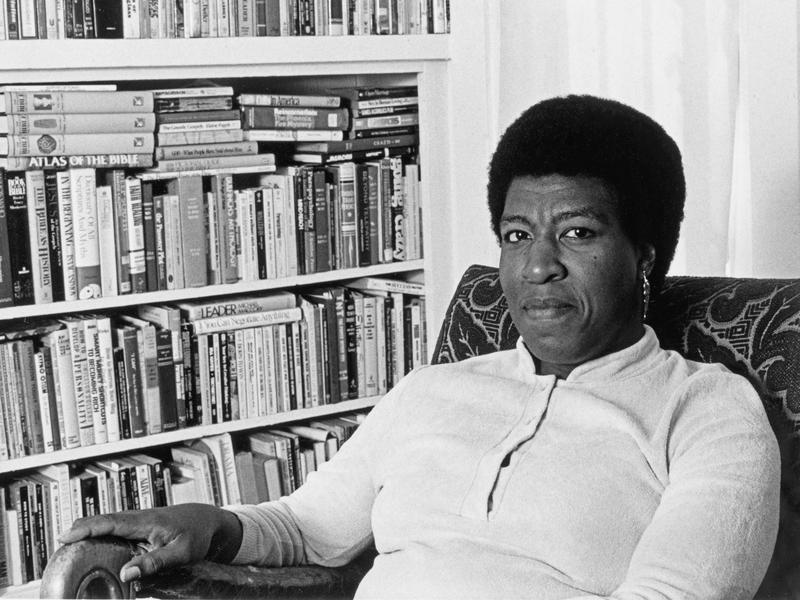 Octavia E. Butler, a Black woman with an afro, is seen seated beside a bookshelf.