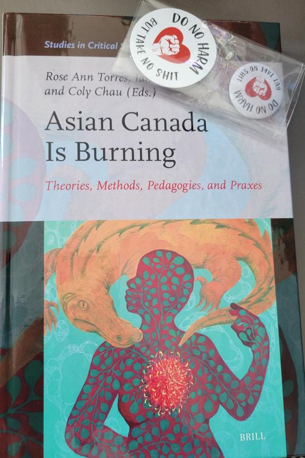 A photo of the latest hard cover anthology with Krystal Kavita Jagoo's essay in it, i.e., Asian Canada is Burning: Theories, Methods, Pedagogies, and Praxes, is seen, on a grey table, with a package of a "DO NO HARM BUT TAKE NO SHIT" sticker and button on top, as created by knezek.art