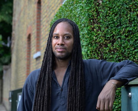 Jason Arday, a Black man with locs is seen, seated outside, with a building with greenery behind him.