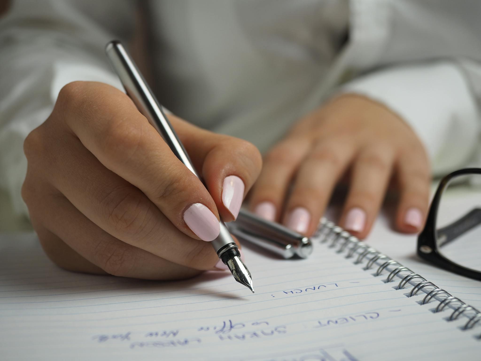 A pair of brown hands with painted nails are seen on a half-written page of an open spiral notebook, in which, a silver pen is held in their right hand, poised to write, with the silver pen cover beside their left hand, and black glasses off to the side.