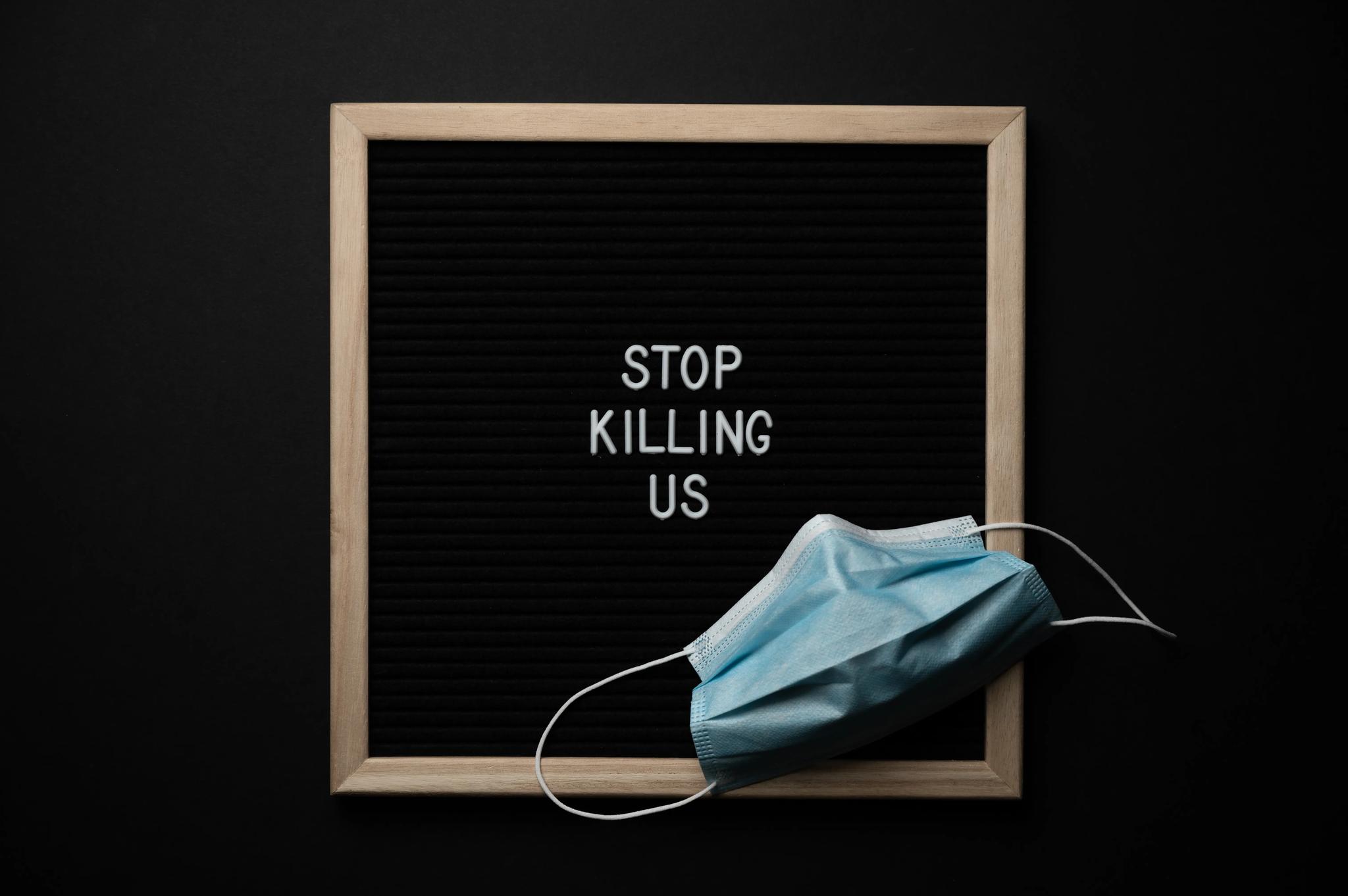 Against a black background, a brown square frame is seen, on which, it states "STOP KILLING US" in white capital letters, with a surgical mask seen in the bottom right corner.