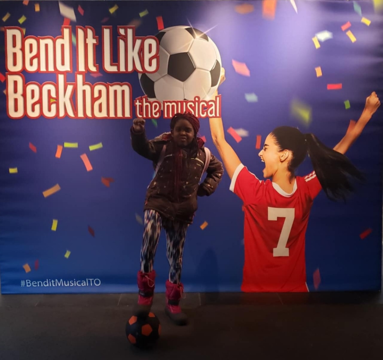 A Black girl is seen posing with a raised fist, and her foot on a soccer ball. In her background, the Bend It Like Beckham poster is seen, which features a brown soccer player in a red uniform.