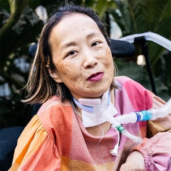 Alice Wong is seen, wearing a colourful shirt, facing the camera, with a slight, possibly mischievous smile on her face.