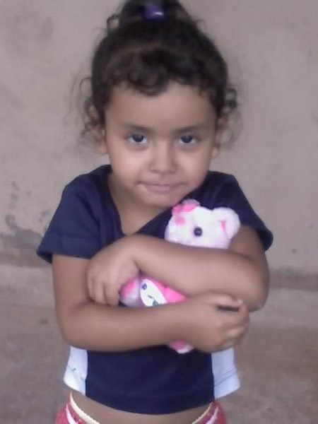 A imagem mostra uma criança parda, vestindo caisa azul e segurando um bichinho de pelúcia cor de rosa