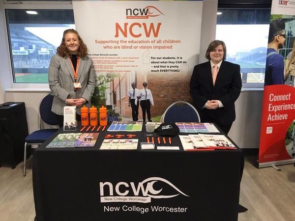 A picture of a stand which bares NCW branding there is a table with a black tablecloth on which has the NCW in white in the centre on the front of the table cloth on top of the table there is a variety of things such as water bottles, information packs, leaflets and pens behind the table to the left is Megan a white women with ginger hair who is wearing a grey blazer and black top in the centre there is a white stand up banner with displays information about NCW on to the right of the picture is Amy (me) who is a white women with dirty blond hair who is wearing a white collared shirt with a orange tie and a black blazer with black trousers.