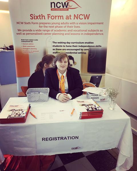 A picture of a stand there is a small table with a plain white table cloth in the centre on the front is a paper sign which reads registration on top of the table is information packs, pens and a small grey box behind the table there is a stand up banner which displays information about NCW Amy a white women with dirty blonde hair is sat on a chair behind the table her arms are on the table and her hands are intertwined she is wearing a white collared shirt with an orange tie and black blazer jacket