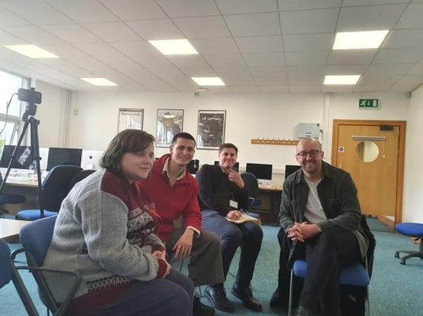 a picture of a room that has a blue carpet and white wallsthere are 4 people sat on chairs from left to right we have Amy a white women with dirty blonde hair who is facing the camera amy have a grey and red jacket on next to Amy is Zac a white man who has short dark hair he is also facing the camera and is wearing a read fleece next to zac is a white man who has a black jumper on he has dark hair and is holding a notebook next to him is Lee Cain who is a white bold man with glasses who is wearing a white tshirt and a brown jacket