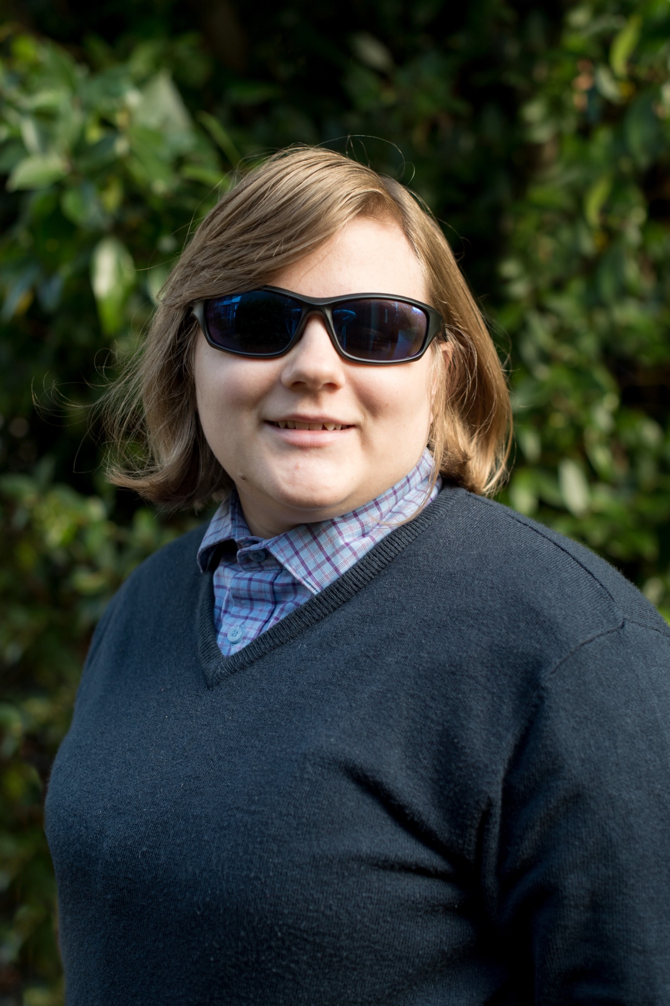 a picture of amy a white women with dirty blonde hair stood against tree leaves amy is smiling towards the camera and is wearing a light blue collared shirt and dark jumper amy is also wearing sunglasses