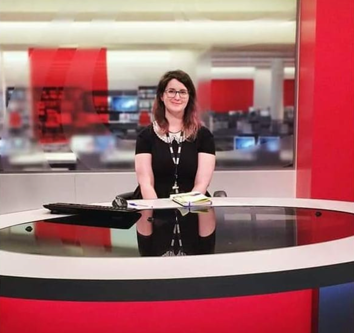 A picture of Maggie Baska, a non-binary/genderqueer journalist, wearing a black and white dress while sitting at the BBC South news set