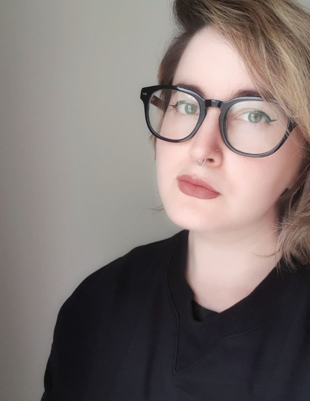 A picture of Maggie Baska, a genderqueer/non-binary journalist, wearing a black top and large glasses as they stare at the camera