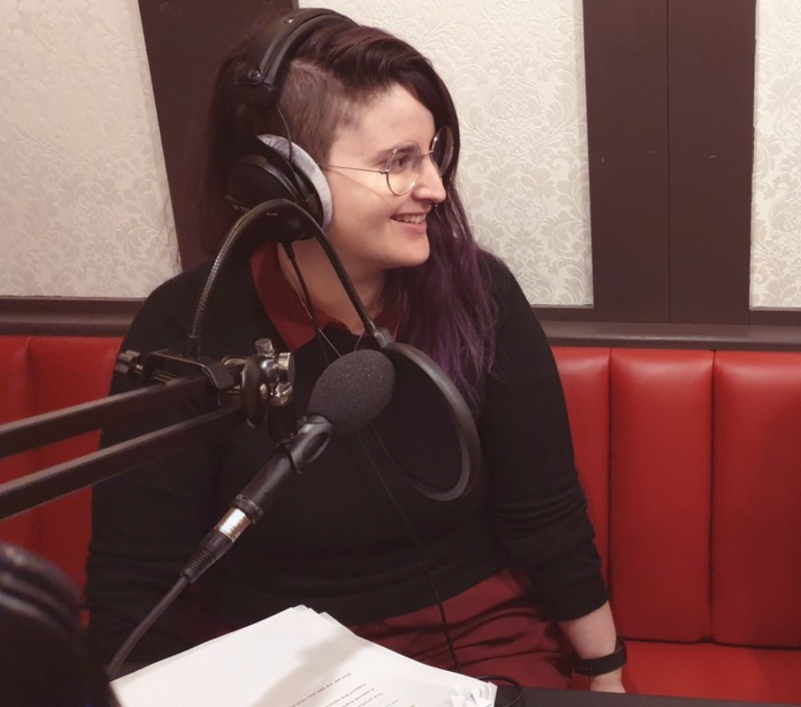 A picture of Maggie Baska, a non-binary/genderqueer journalist, wearing a dark top and red dress as they record a podcast