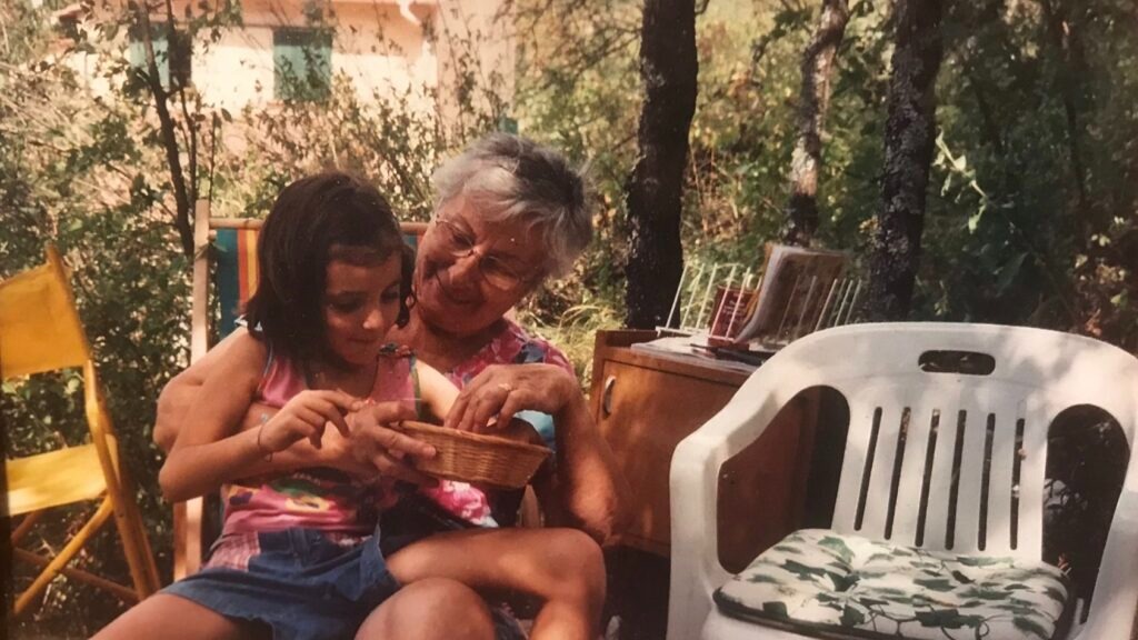 Caterina is sitting on her Nonna Nevina's lap while they are eating berries from a weave basket in the countryside. Caterina is sitting on her Nonna Nevina's lap while they are eating berries from a weave basket in the countryside.