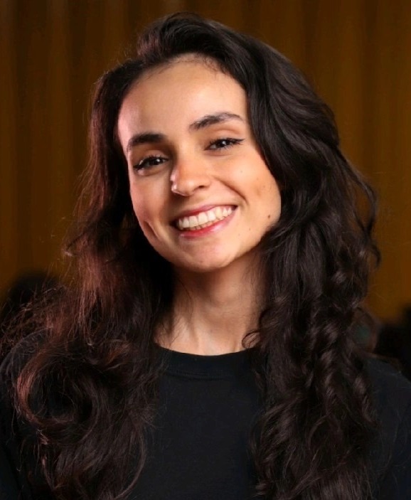 Fotografia em retrato de Barbara Viotto. Ela é uma mulher jovem de pele clara. Ela tem cabelo castanho, ondulado, com comprimento para baixo dos seios. Seus olhos e sobrancelhas também são castanhos. Barbara olha para a câmera, com a cabela levemente inclinada para a esquerda, abre um grande sorriso.